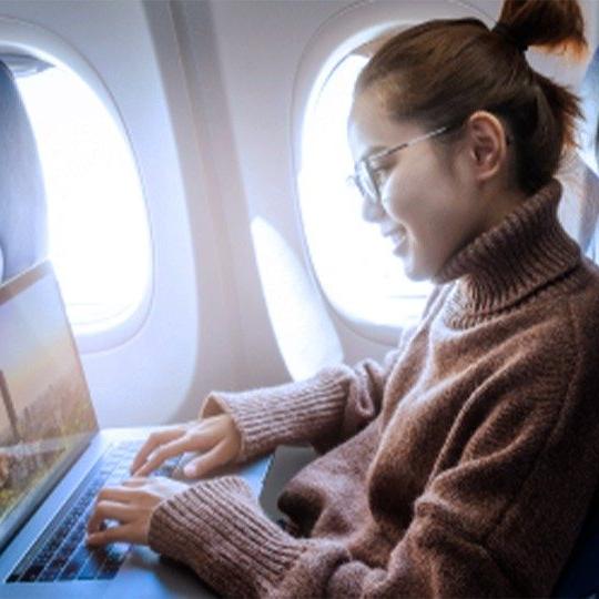 一名戴眼镜、穿着棕色毛衣的女子在飞机上用笔记本电脑通过机上WiFi工作